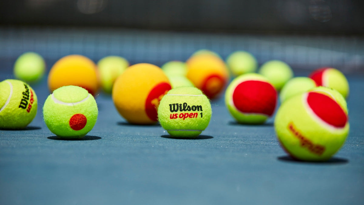 Pelotas de tenis sobre una pista dura: una amarilla normal en el centro y, alrededor, pelotas de iniciación rojas y naranjas.