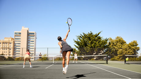 Jugadora de tenis de espaldas sacando en una pista dura, con la red y otros tres jugadores al fondo.